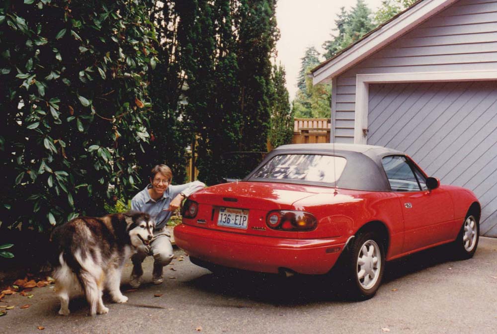 A person crouching beside a large dog in front of a red convertible parked next to a house, with green trees and shrubs in the background.