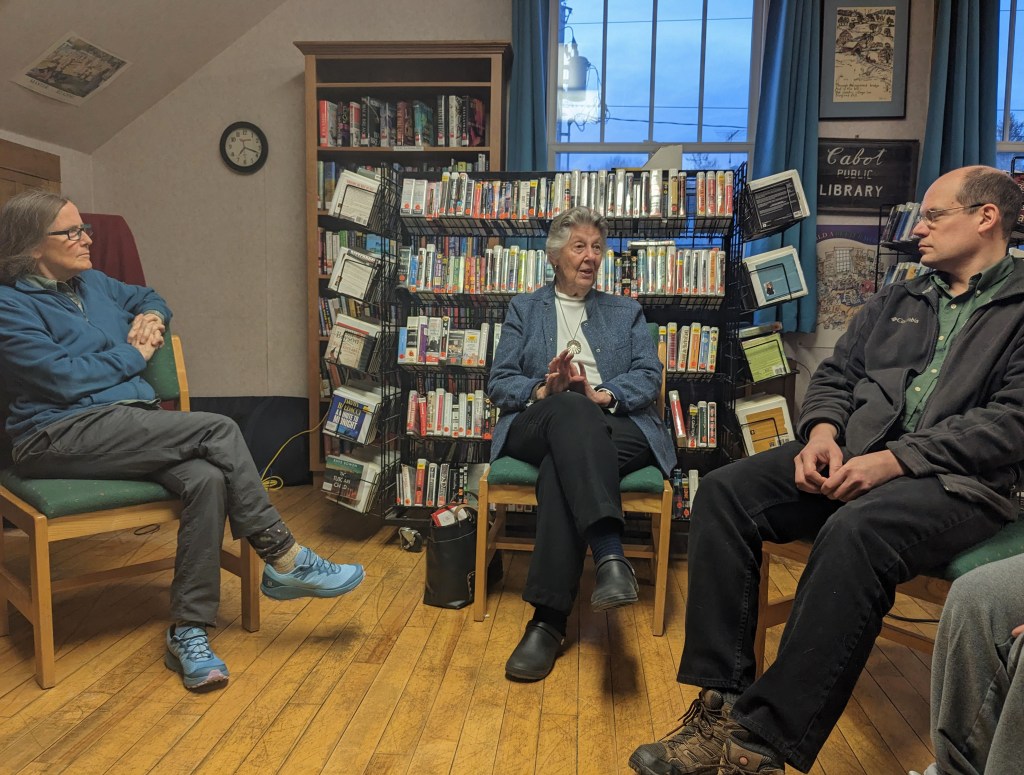 A discussion taking place in a small library, featuring three people seated in chairs. One person is speaking while the others listen attentively, surrounded by shelves filled with books and media.